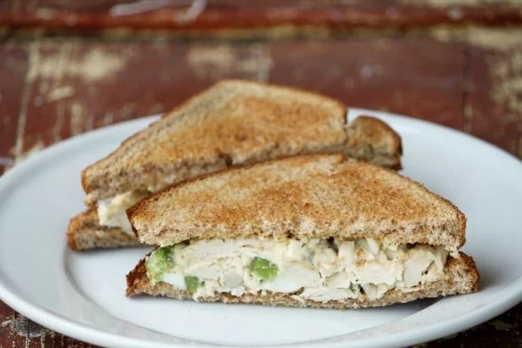 Creamy chicken salad sandwich garnished with fresh herbs on a wooden cutting board.
